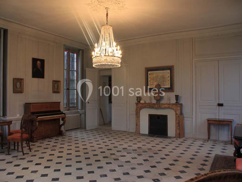 Salon ancien avec un lustre en cristal, cheminée en marbre, piano droit et sol en carreaux noirs et blancs.