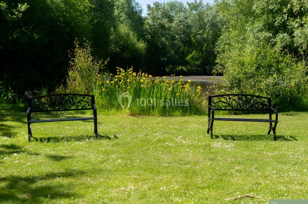 Deux bancs en métal noir sur une pelouse, face à un étang entouré de végétation et de roseaux.