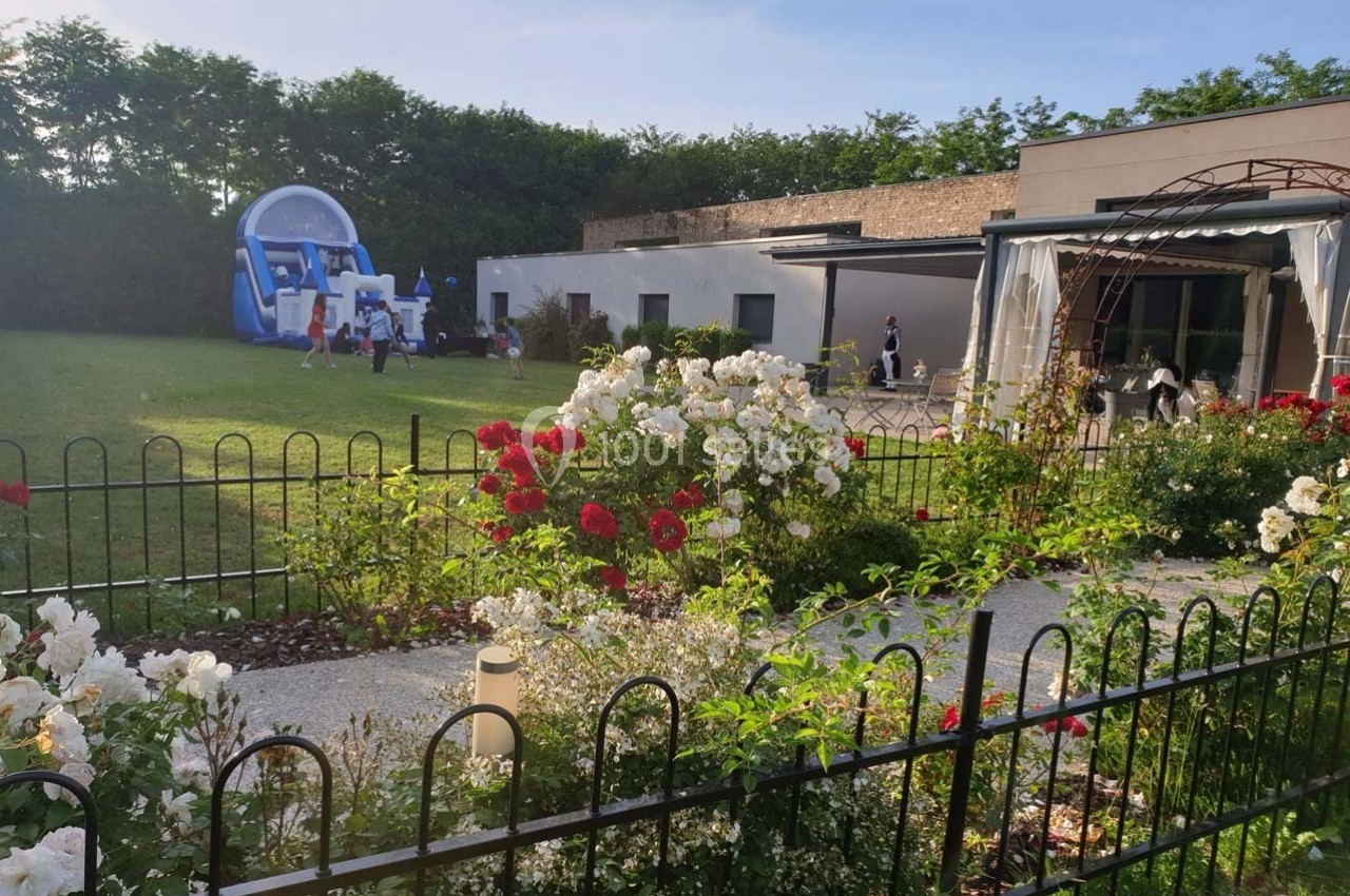 Jardin fleuri avec rosiers, pelouse, structure gonflable en arrière-plan et bâtiment moderne à droite.