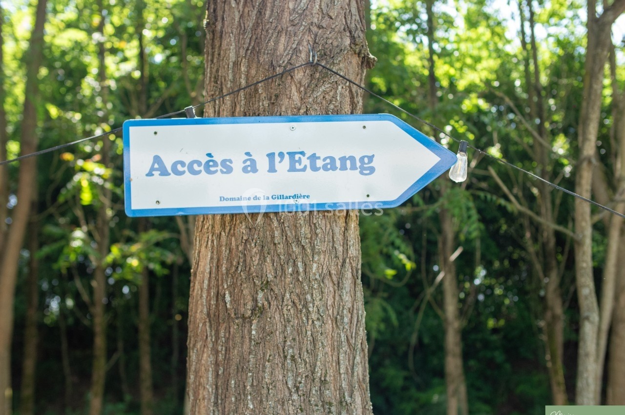Panneau indiquant ’Accès à l'Étang’ fixé à un arbre, avec une forêt en arrière-plan.