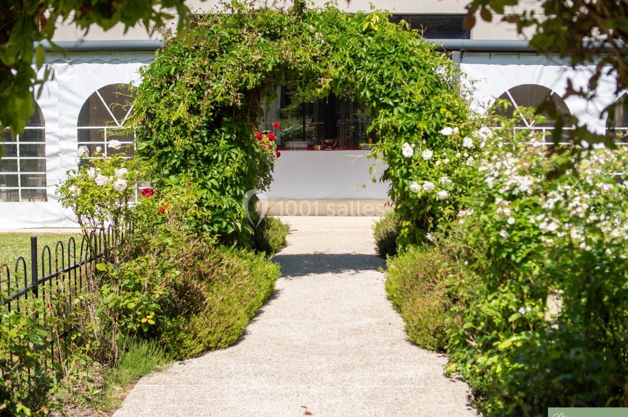 Allée bordée de verdure menant à une arche végétale devant un bâtiment avec des fenêtres en arc.