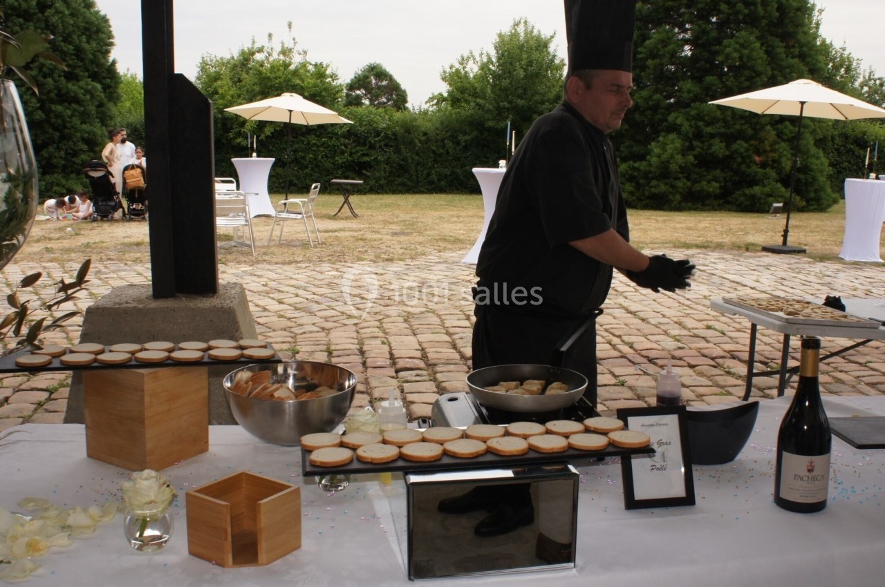Un chef prépare des amuse-bouches sur une table dressée en extérieur, avec des parasols et un jardin en arrière-plan.