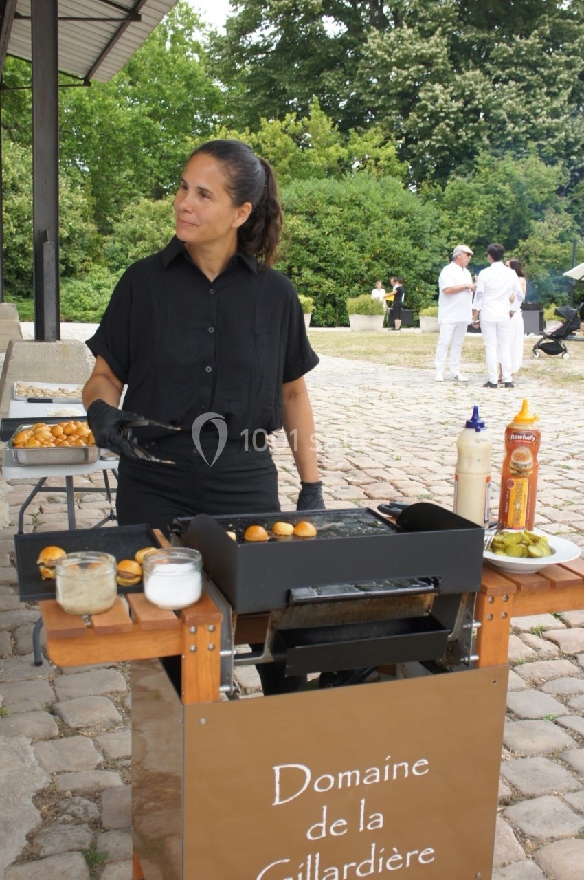 Femme en tenue noire cuisinant des aliments sur une plancha devant un stand en extérieur, avec des arbres en arrière-plan.