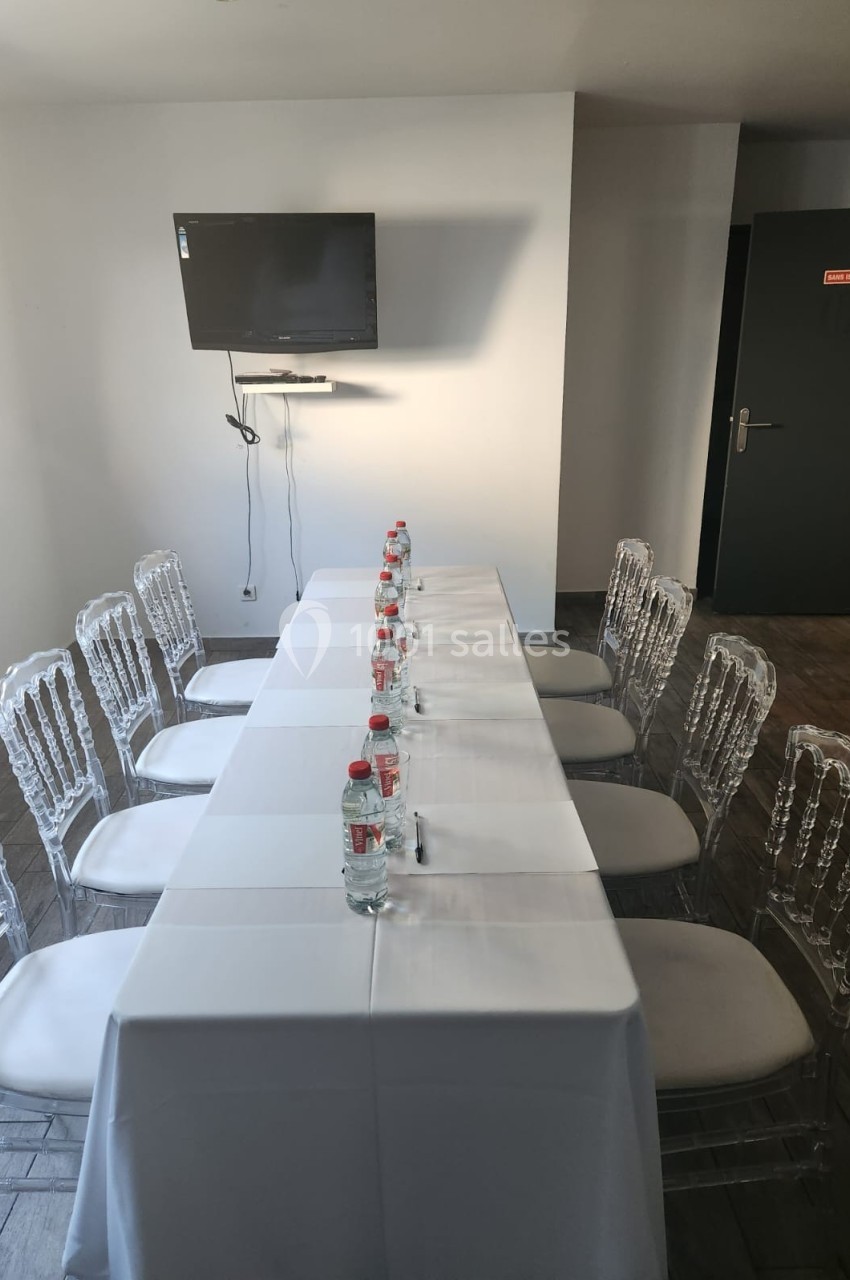 Salle de réunion avec une table rectangulaire couverte d'une nappe blanche, chaises transparentes et écran mural.