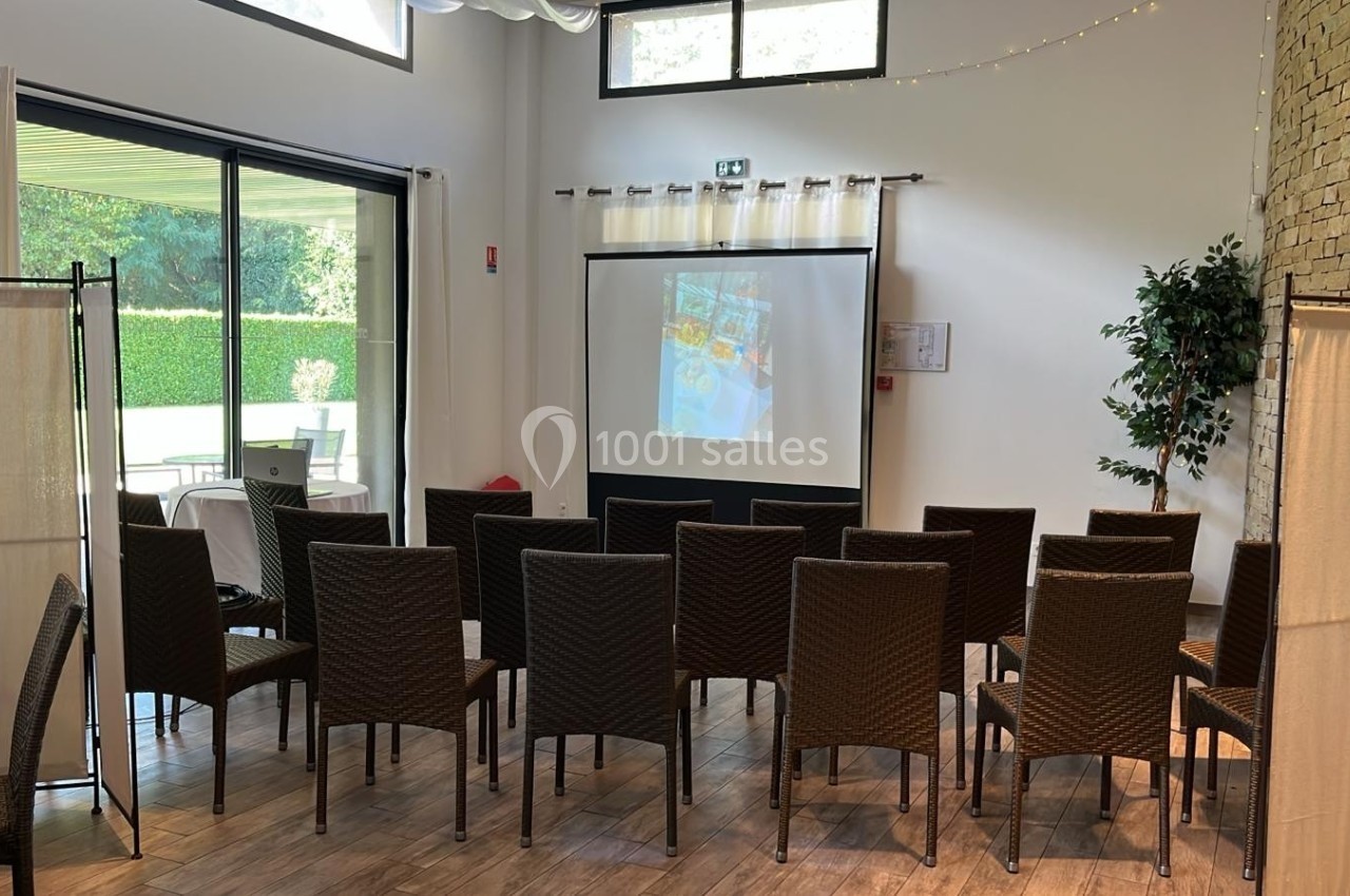 Salle aménagée avec des chaises en rotin face à un écran de projection, éclairée par la lumière naturelle d'une baie vitrée.