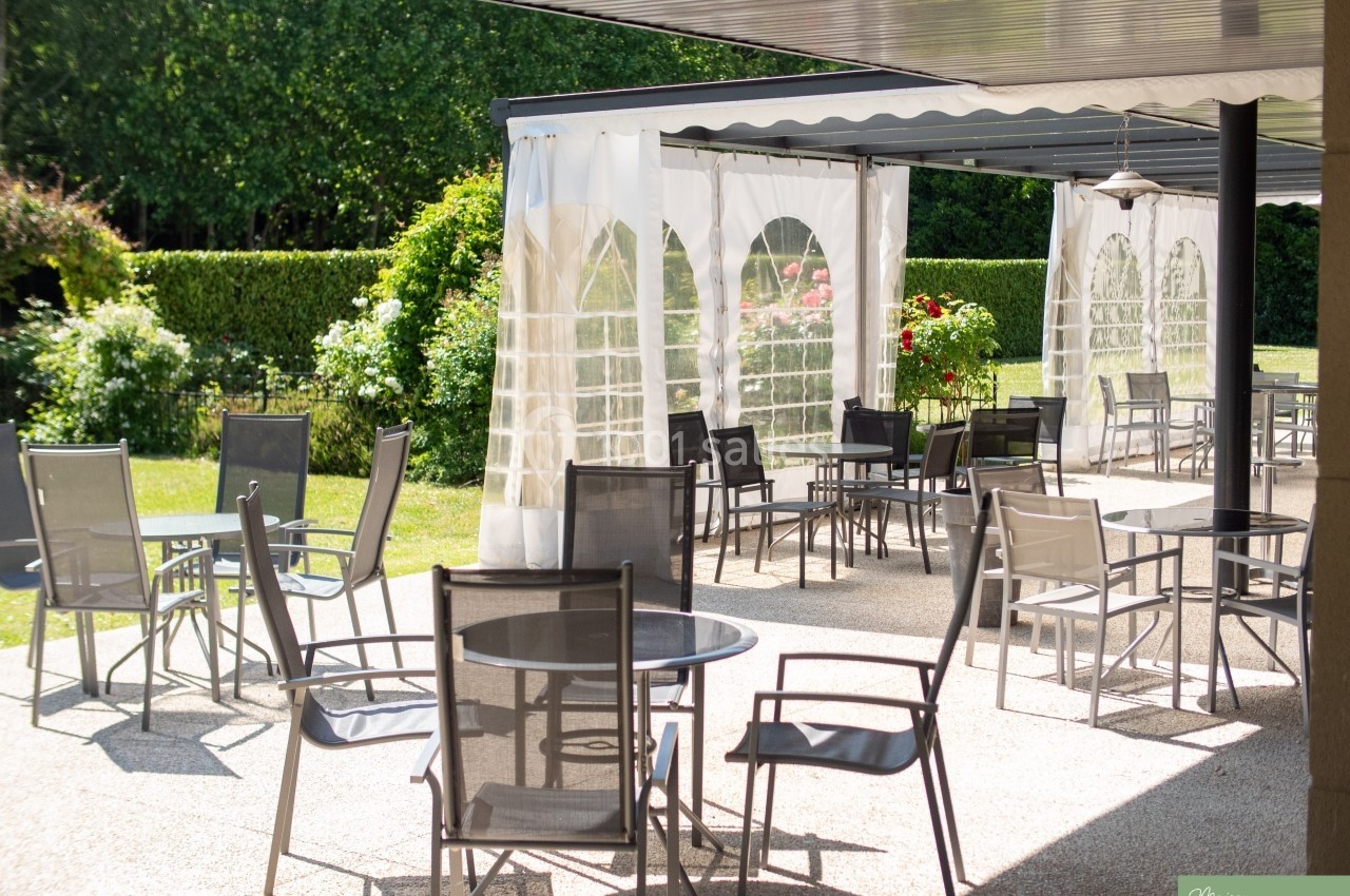 Terrasse extérieure avec tables et chaises en métal, partiellement ombragée par une pergola et entourée de verdure.