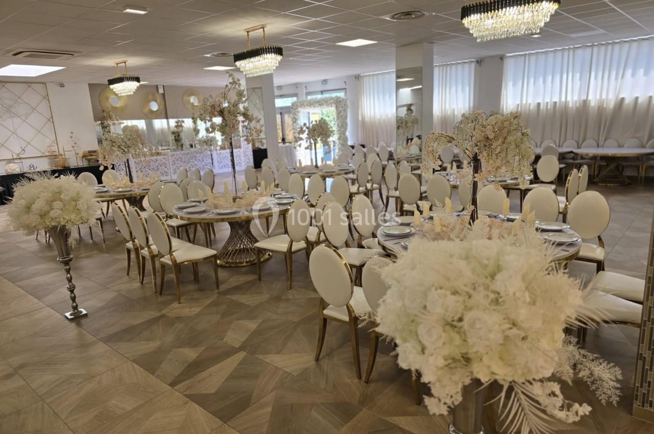 Salle de réception élégamment décorée avec des tables rondes, des chaises blanches et des compositions florales blanches.