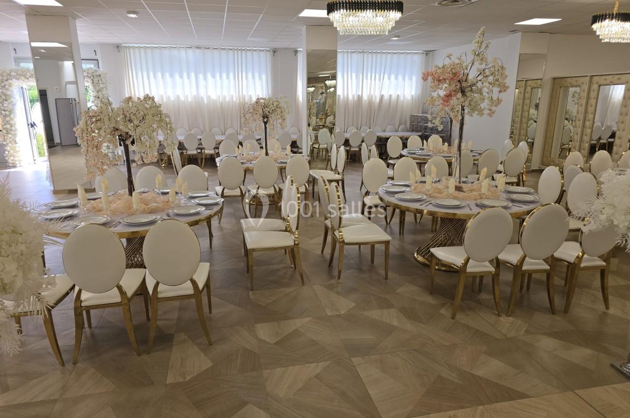 Salle de réception élégante avec tables rondes décorées, chaises blanches et dorées, et arrangements floraux.