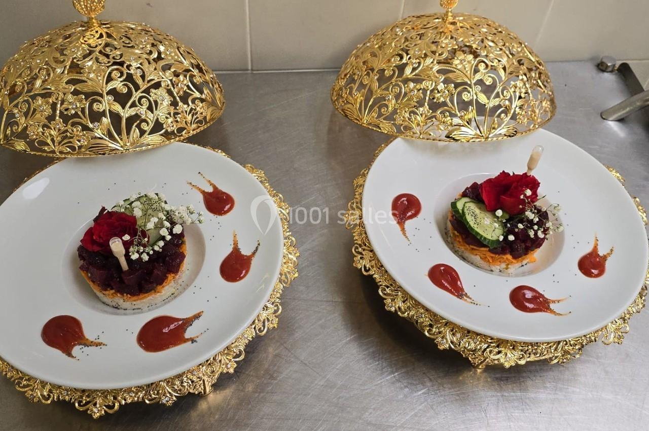 Deux assiettes décorées avec des plats raffinés sous des cloches dorées, présentées sur une table en inox.