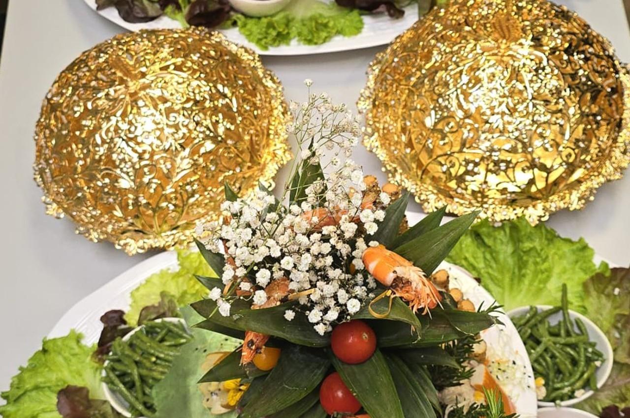 Plateau décoratif avec légumes, crevettes et fleurs comestibles, entouré de cloches dorées et de feuilles de salade.