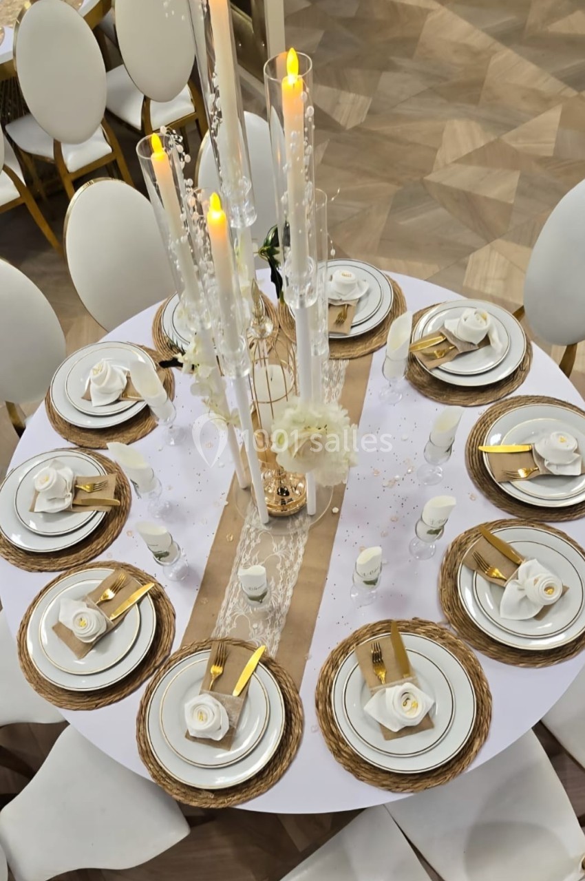 Table ronde élégamment dressée avec vaisselle blanche, serviettes dorées, bougies et décorations florales blanches.