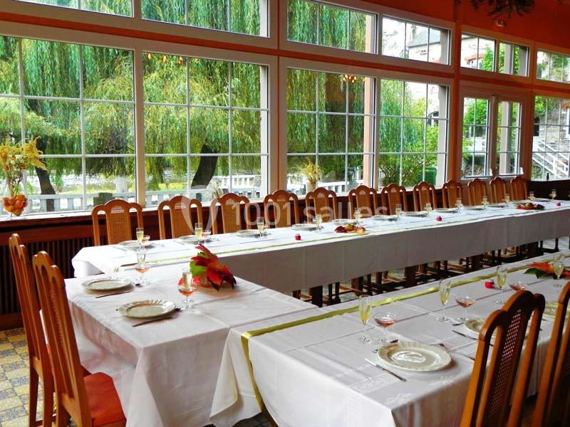 Salle lumineuse avec de grandes baies vitrées, tables dressées avec nappes blanches, verres et assiettes.