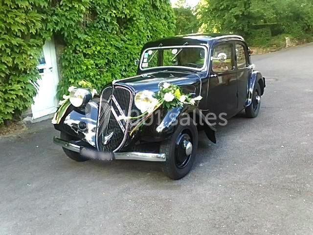 Voiture ancienne noire décorée de fleurs blanches, garée sur une allée asphaltée devant un mur couvert de lierre.