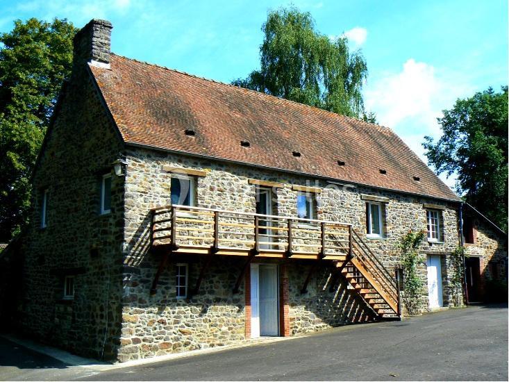 Maison en pierre avec toit en tuiles, balcon en bois et escalier extérieur, entourée d'arbres et d'un ciel dégagé.