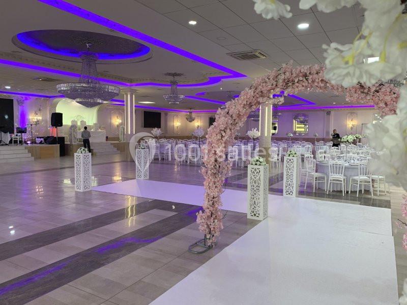 Salle de réception élégante avec tables dressées, arche florale rose et éclairage violet au plafond.