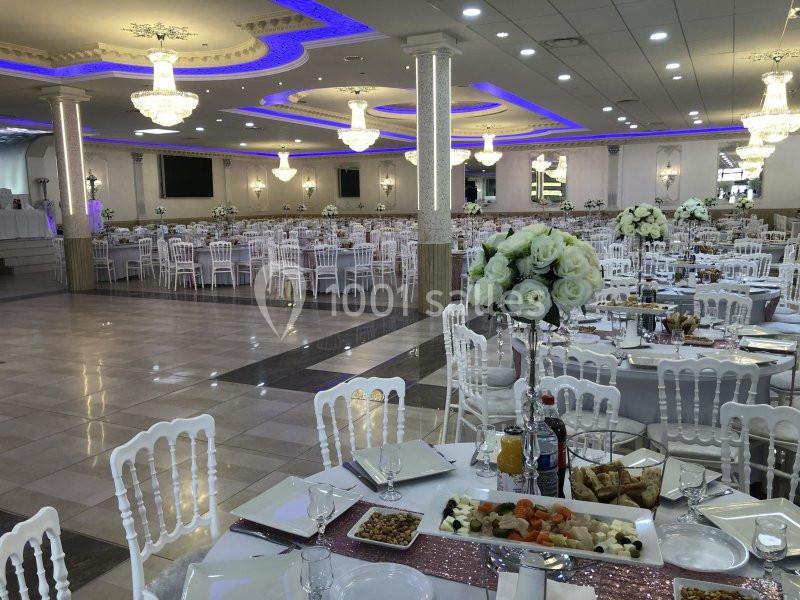 Salle de réception élégante avec tables dressées, chaises blanches et décorations florales sous des lustres lumineux.