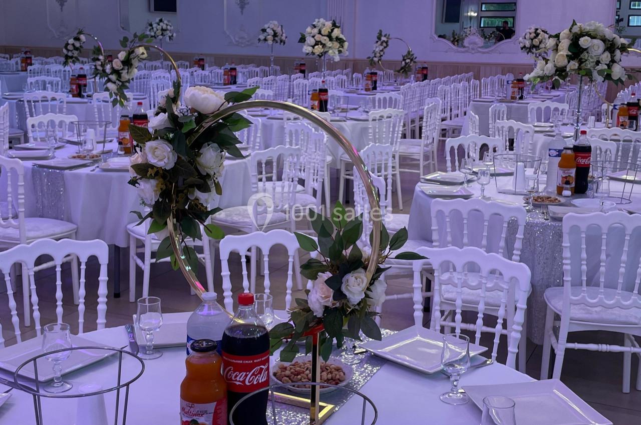 Salle de réception décorée avec des tables rondes, nappes blanches, chaises assorties et centres de table fleuris.