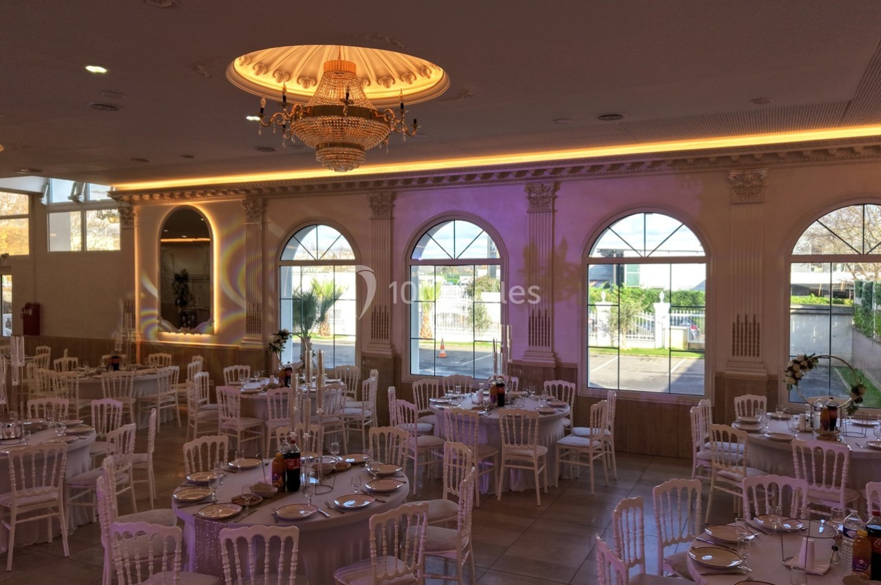 Salle de réception élégante avec tables dressées, chaises blanches et grandes fenêtres laissant entrer la lumière naturelle.