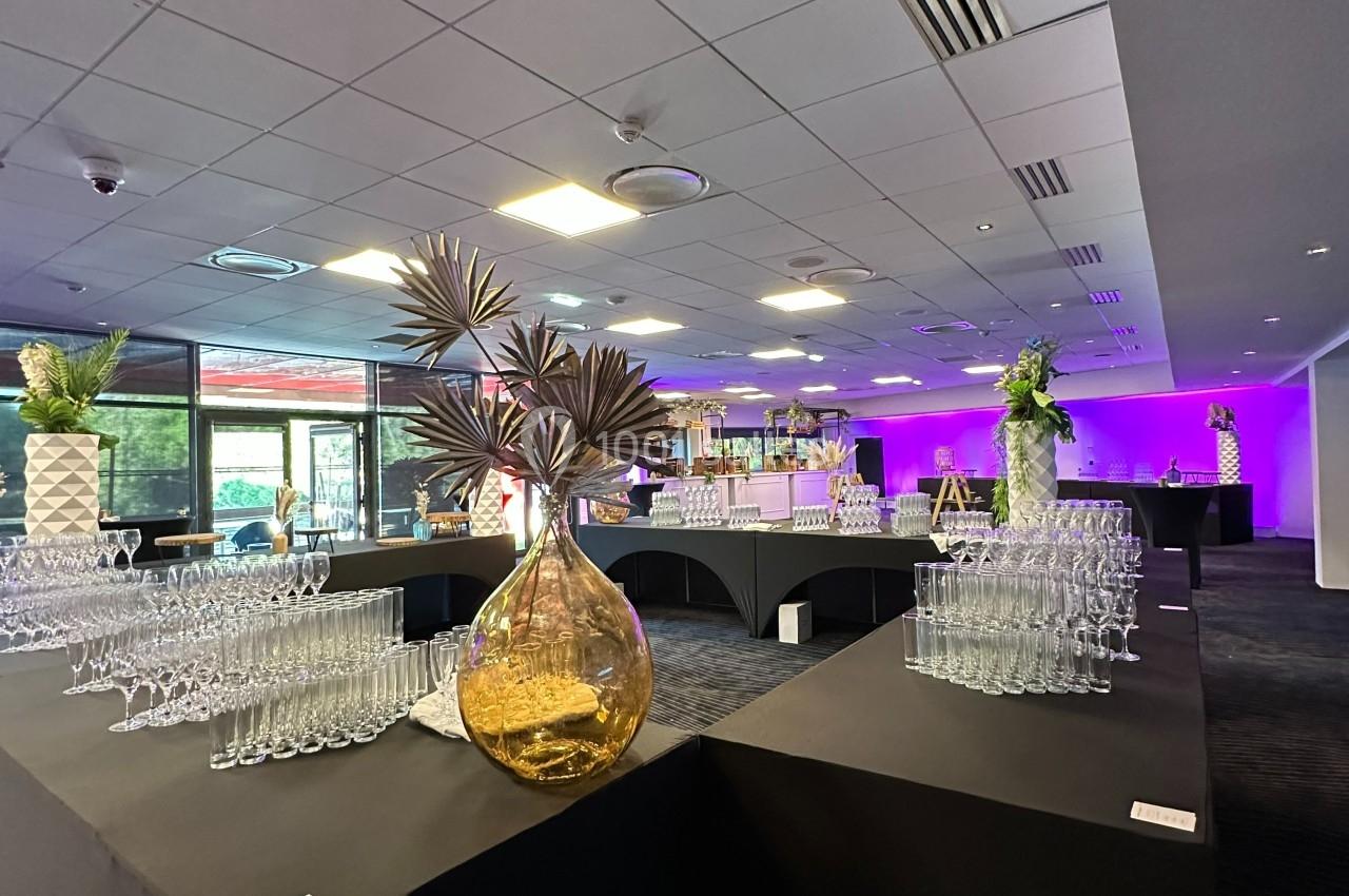 Salle de réception avec tables noires, verres disposés, décoration florale et éclairage violet en arrière-plan.