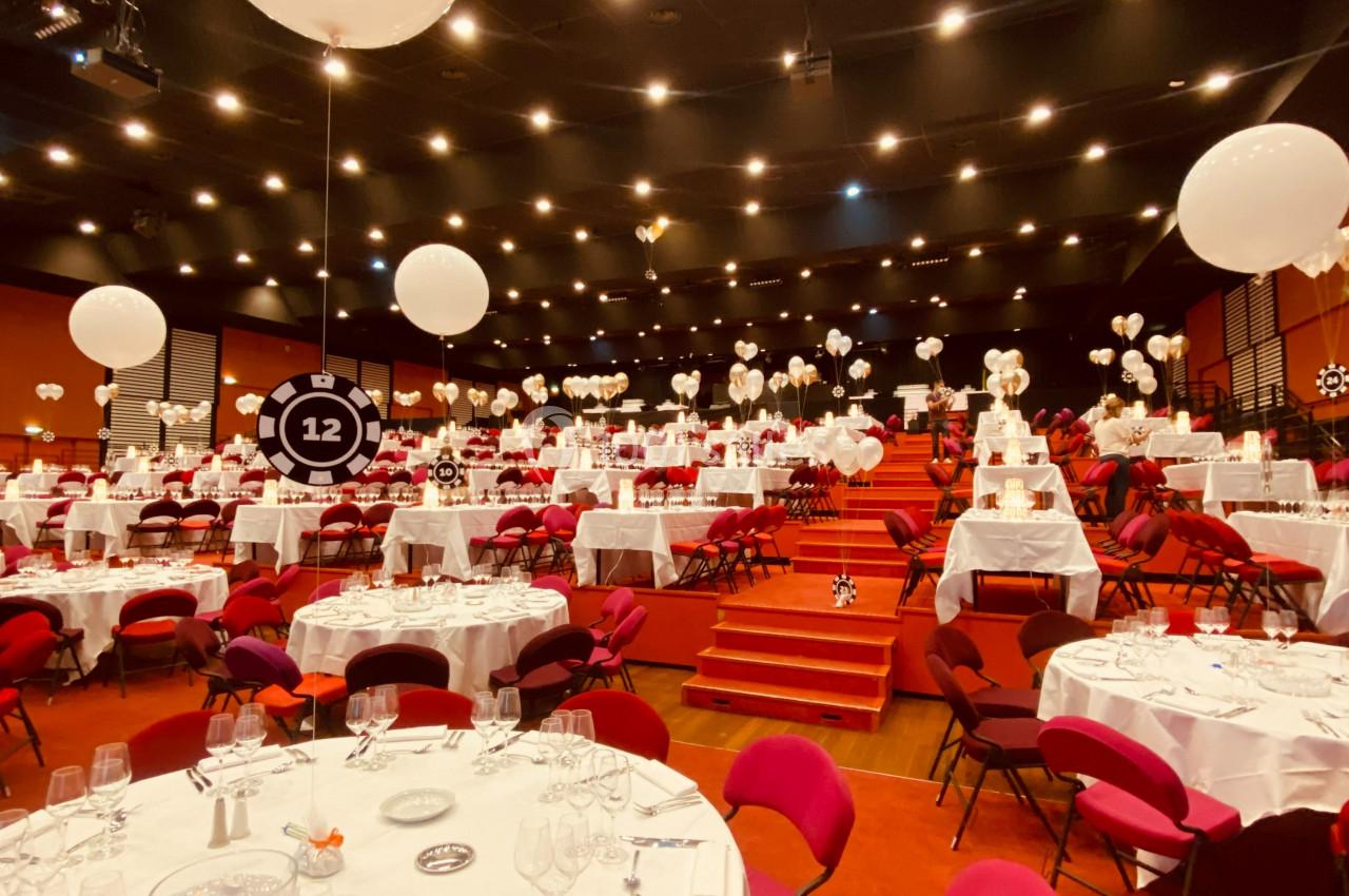 Salle de réception décorée avec des ballons blancs, des tables rondes dressées et des chaises rouges.