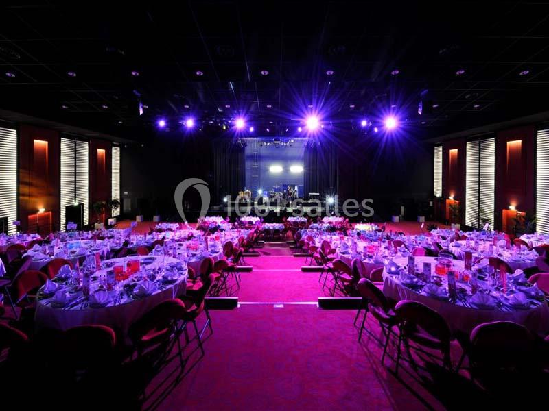 Salle de réception avec tables rondes dressées et éclairage violet, scène en fond avec équipement musical.