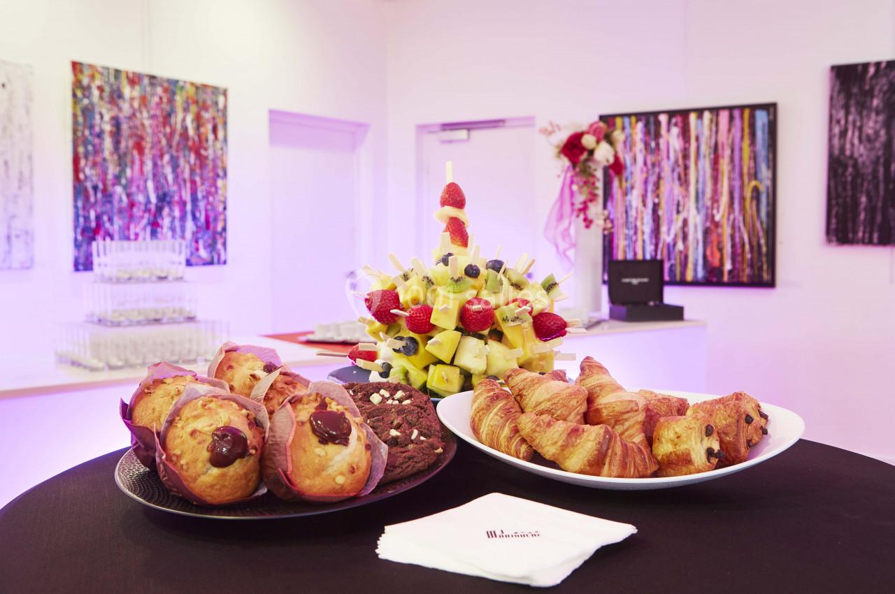 Assortiment de viennoiseries, muffins et brochettes de fruits sur une table dans une salle décorée de tableaux colorés.