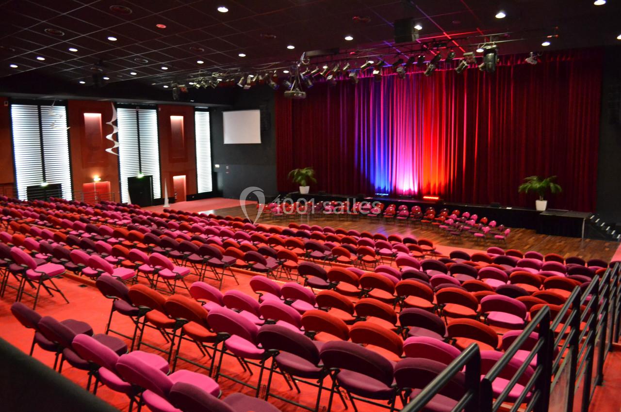 Salle de spectacle avec rangées de sièges rouges et roses, scène éclairée par des lumières colorées et rideaux rouges.