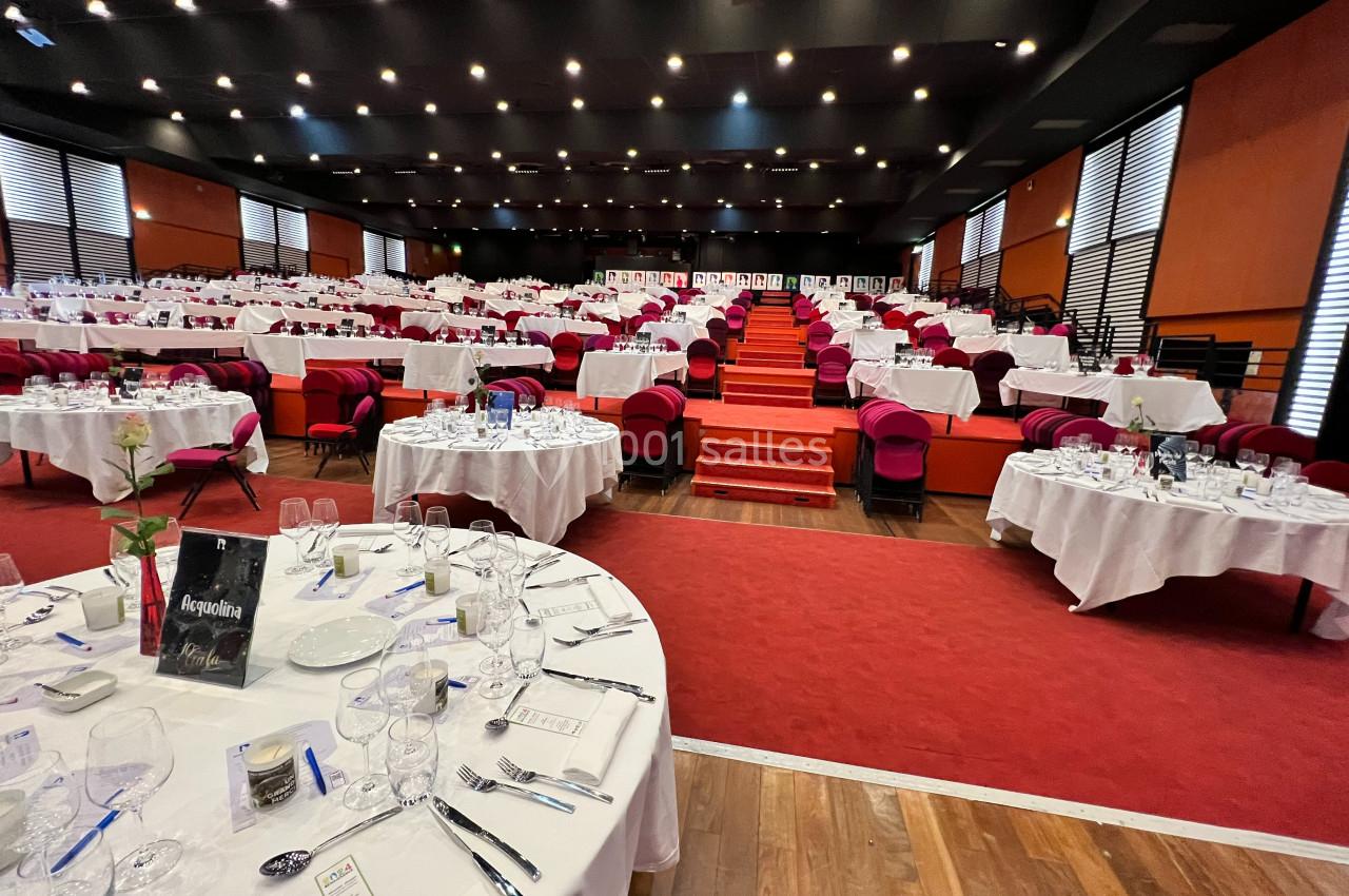 Salle de réception avec tables rondes dressées, nappes blanches, vaisselle élégante et sièges rouges disposés en gradins.