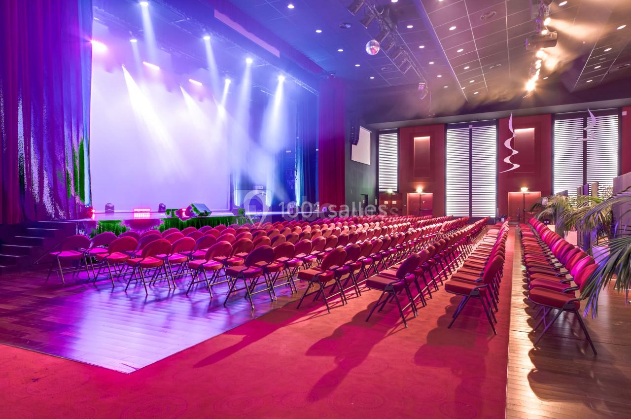 Salle de spectacle avec rangées de sièges rouges, scène éclairée par des projecteurs et rideaux ouverts.