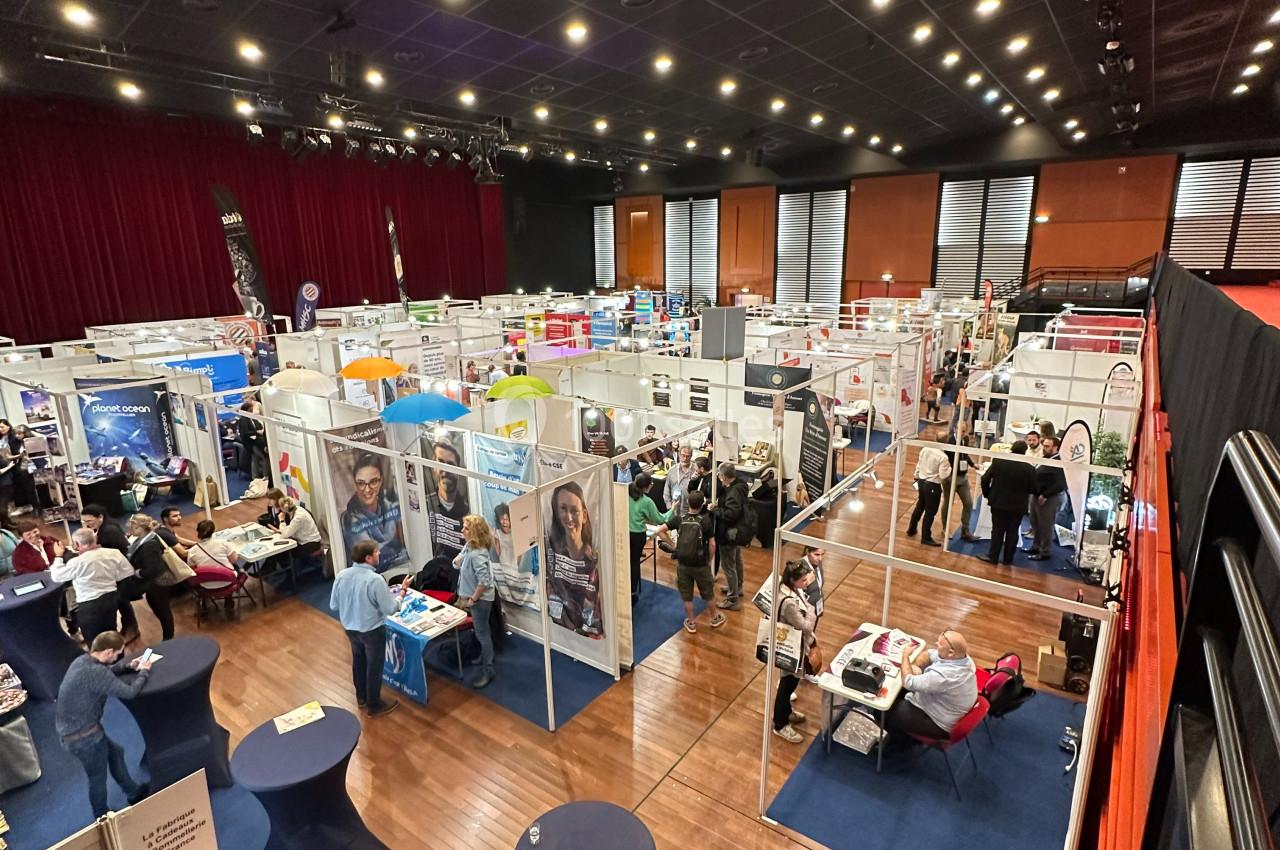 Vue d'ensemble d'un salon professionnel avec des stands, des visiteurs et une scène dans une grande salle éclairée.