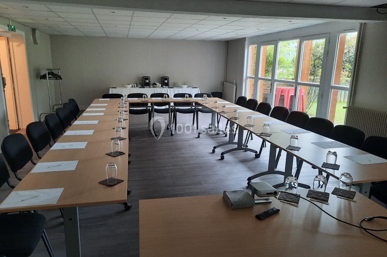 Salle de réunion lumineuse avec tables disposées en U, chaises, verres, papiers et vue sur un jardin à travers de grandes…