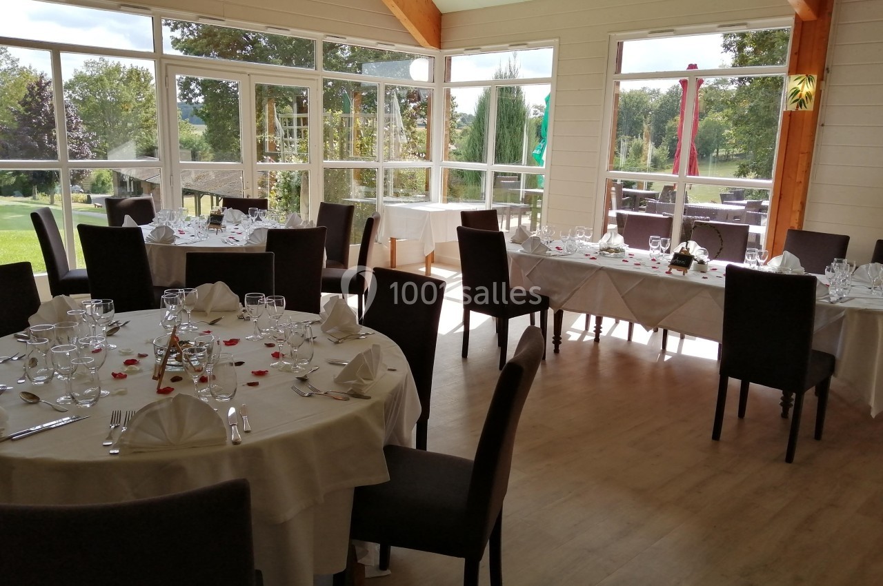 Salle lumineuse avec grandes baies vitrées, tables rondes et rectangulaires dressées pour un repas, vue sur un jardin.
