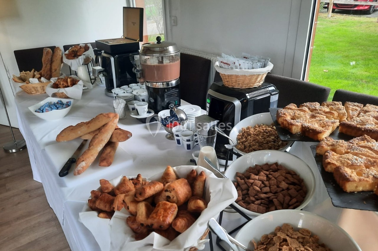 Buffet de petit-déjeuner avec viennoiseries, pains, céréales, boissons chaudes et condiments sur une table blanche.
