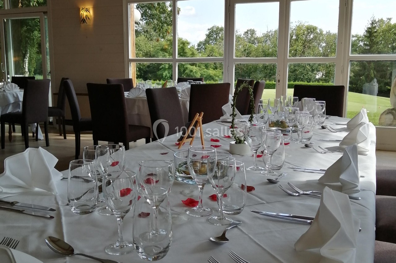 Table élégamment dressée avec nappes blanches, verres, couverts et décorations florales, dans une salle lumineuse avec vue…