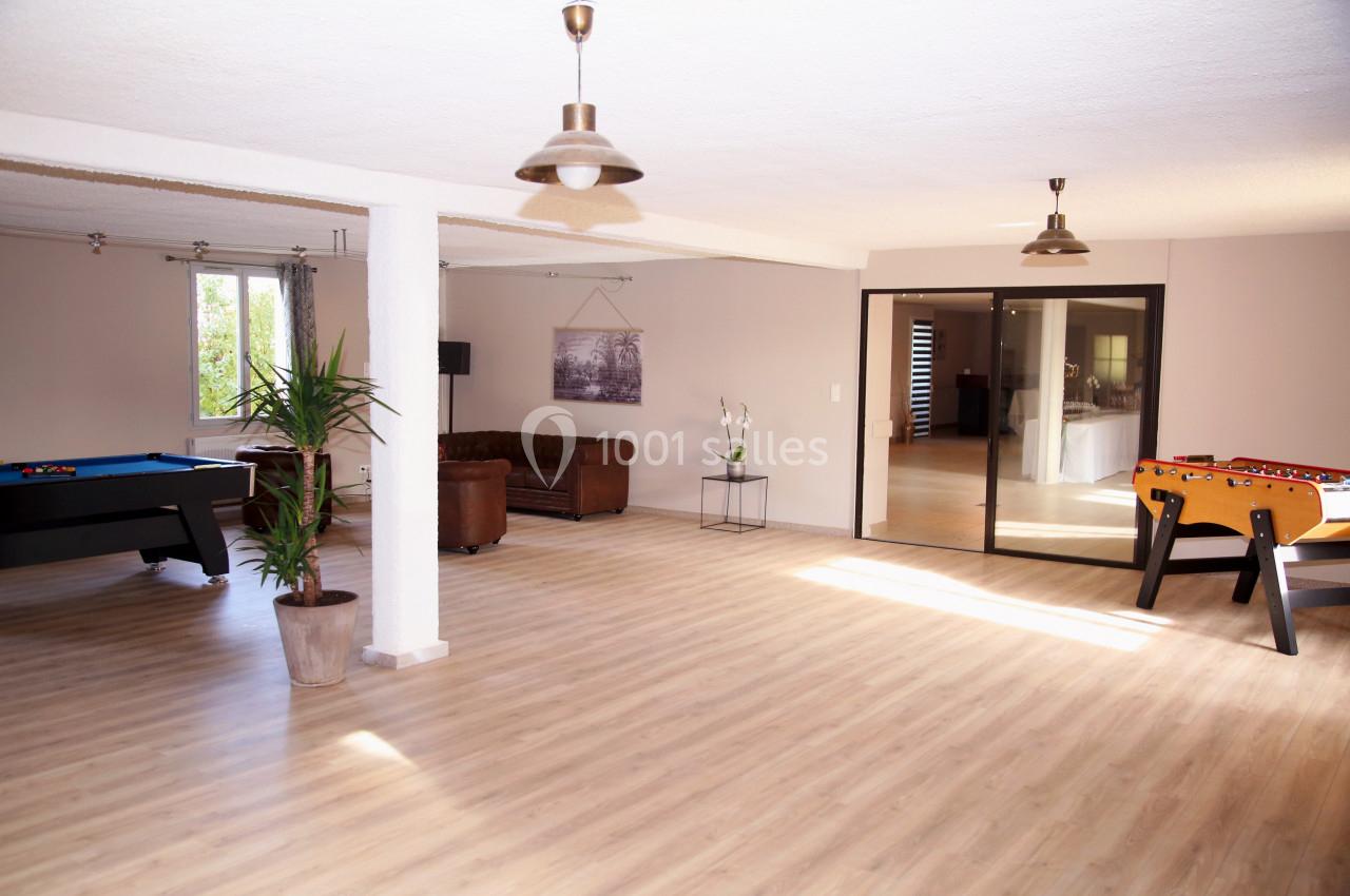 Salle spacieuse avec parquet clair, billard, baby-foot, canapé en cuir, plantes et grande baie vitrée.