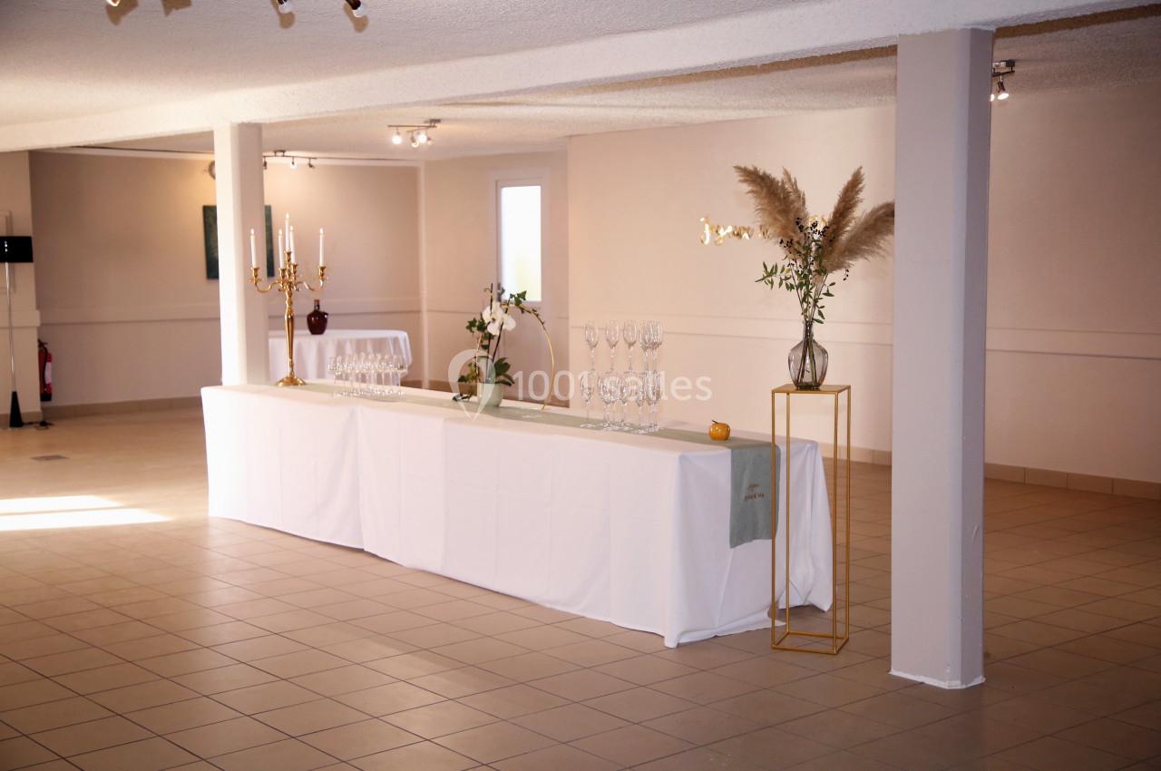 Table dressée avec nappes blanches, chandeliers, verres et décorations florales dans une salle lumineuse au sol carrelé.