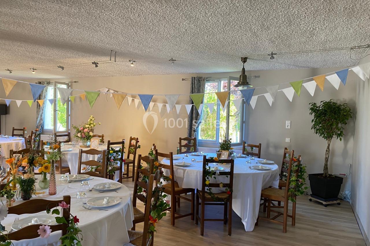Salle décorée pour un événement, avec tables rondes dressées, guirlandes colorées et plantes en arrière-plan.