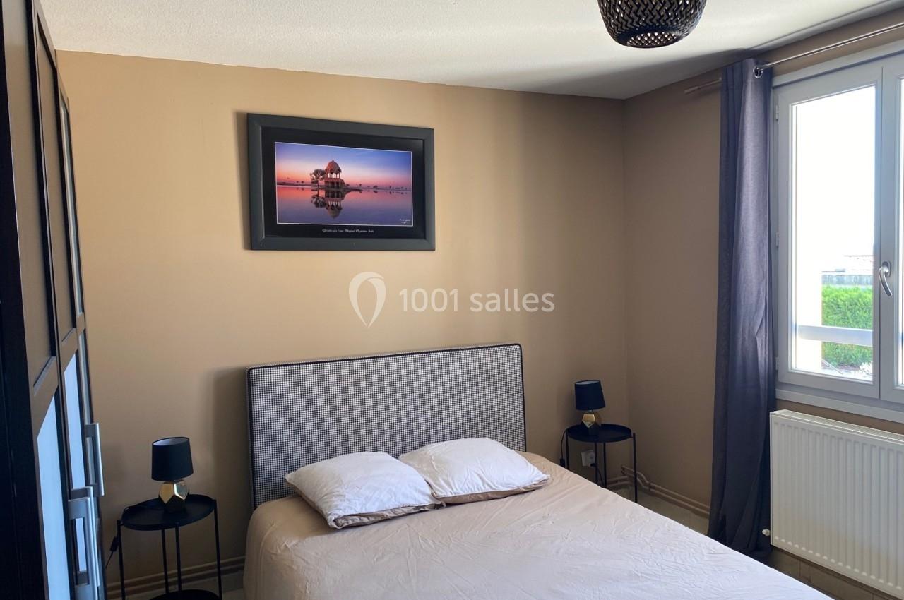 Chambre avec lit double, murs beige, cadre photo au mur, deux tables de chevet et une fenêtre avec rideaux gris.