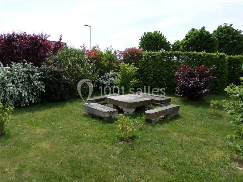 Table en pierre entourée de bancs dans un jardin verdoyant avec des haies et des arbustes fleuris.