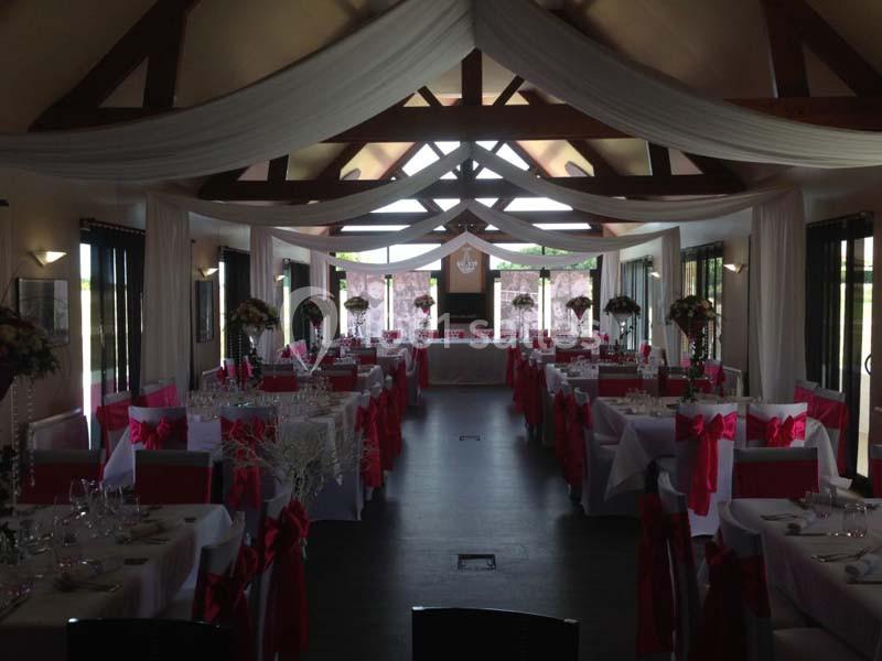 Salle de réception décorée pour un événement, avec nappes blanches, chaises ornées de nœuds rouges et tables fleuries.