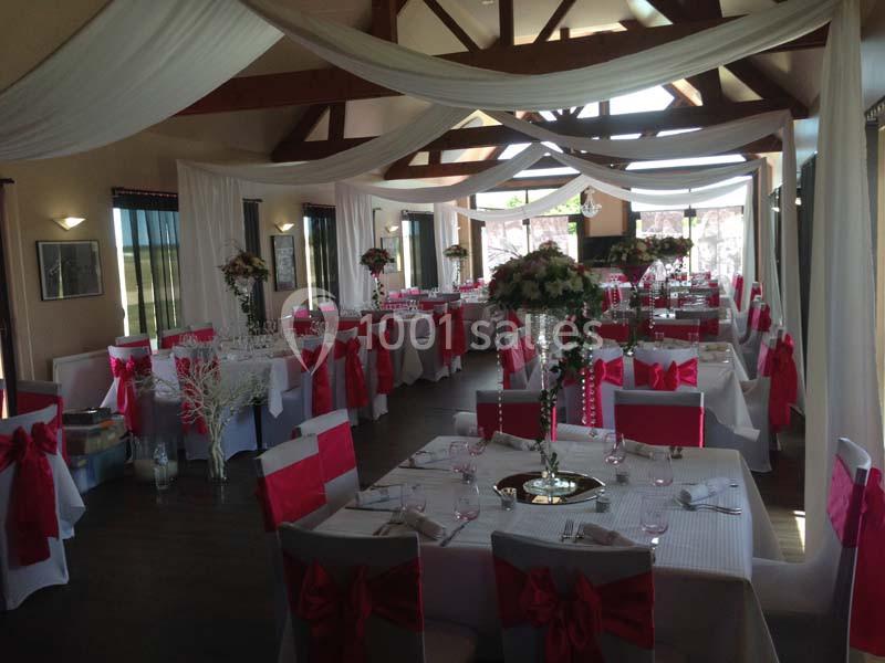 Salle de réception décorée pour un événement, avec nappes blanches, chaises ornées de nœuds rouges et arrangements floraux.