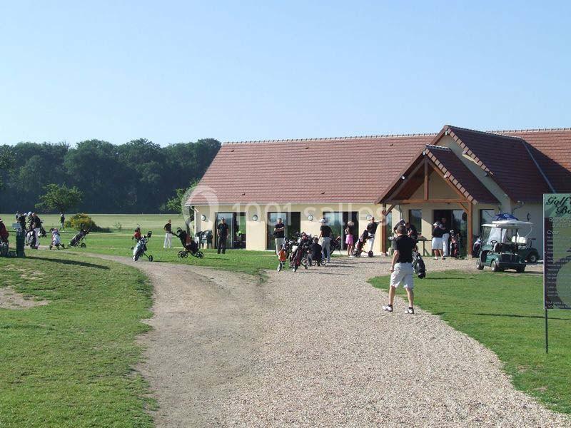 Des golfeurs et visiteurs se rassemblent près d'un bâtiment avec toit en tuiles, entouré de pelouses et d'arbres.