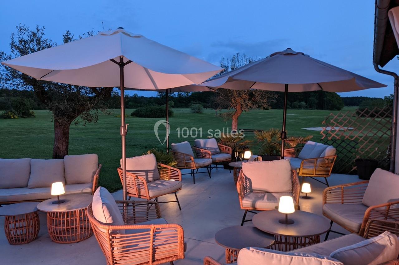 Terrasse aménagée avec des fauteuils en rotin, des parasols et des lampes, donnant sur un jardin au crépuscule.