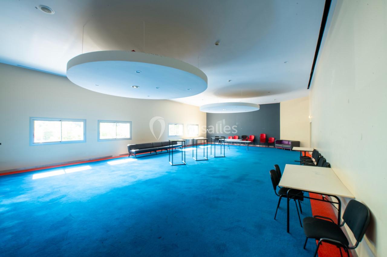 Salle lumineuse avec moquette bleue, tables, chaises, canapés et grandes suspensions rondes au plafond.