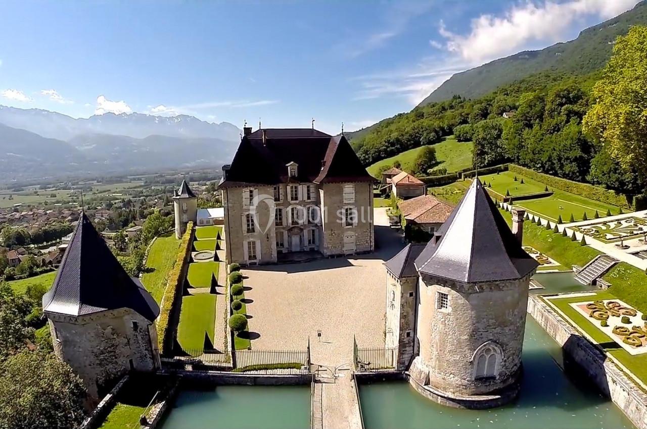 Vue aérienne d'un château entouré de tours, d'un pont et de jardins, situé dans un paysage vallonné avec des montagnes.