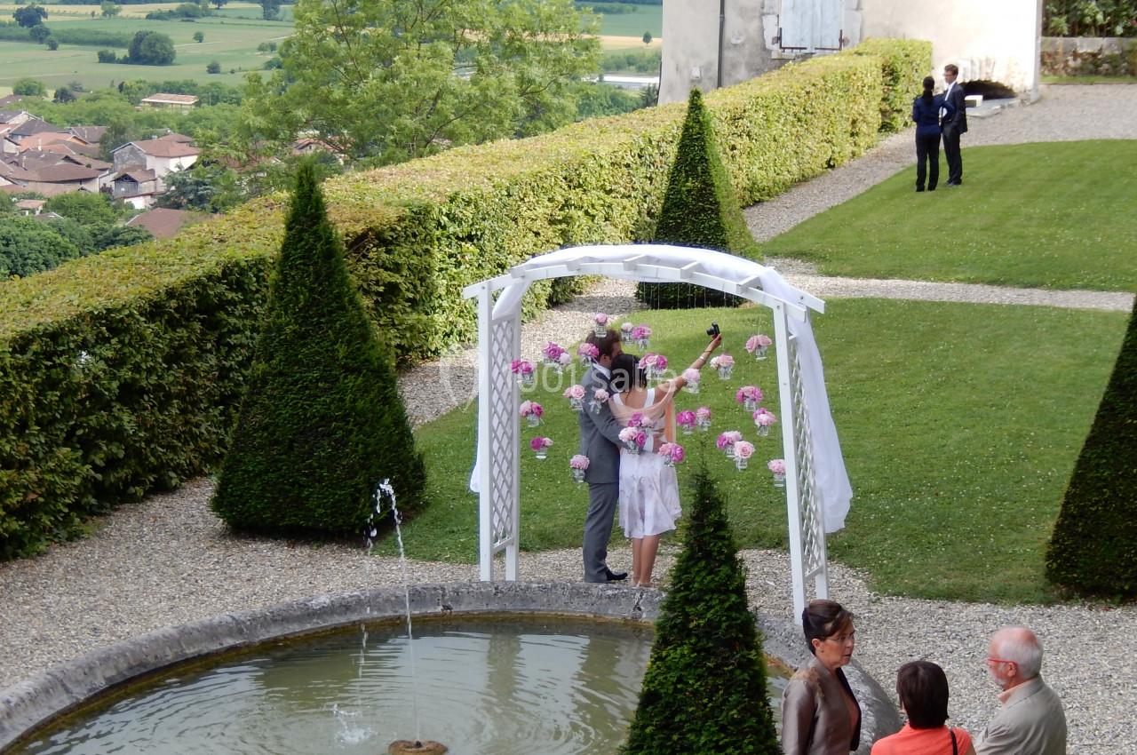 Un couple célèbre son mariage sous une arche décorée de fleurs, dans un jardin avec fontaine et vue sur la campagne.