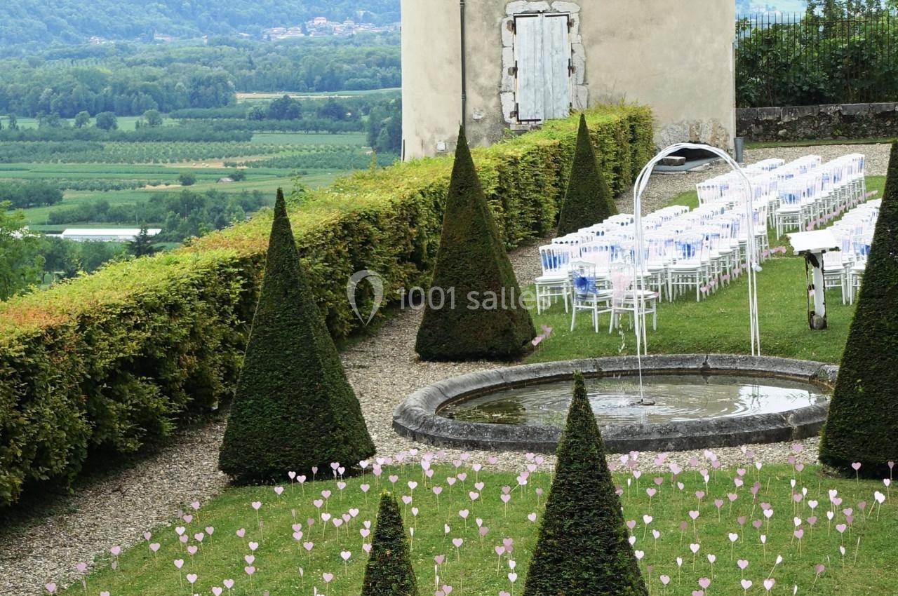 Jardin avec haies taillées en cônes, fontaine centrale et rangées de chaises blanches disposées pour un événement.