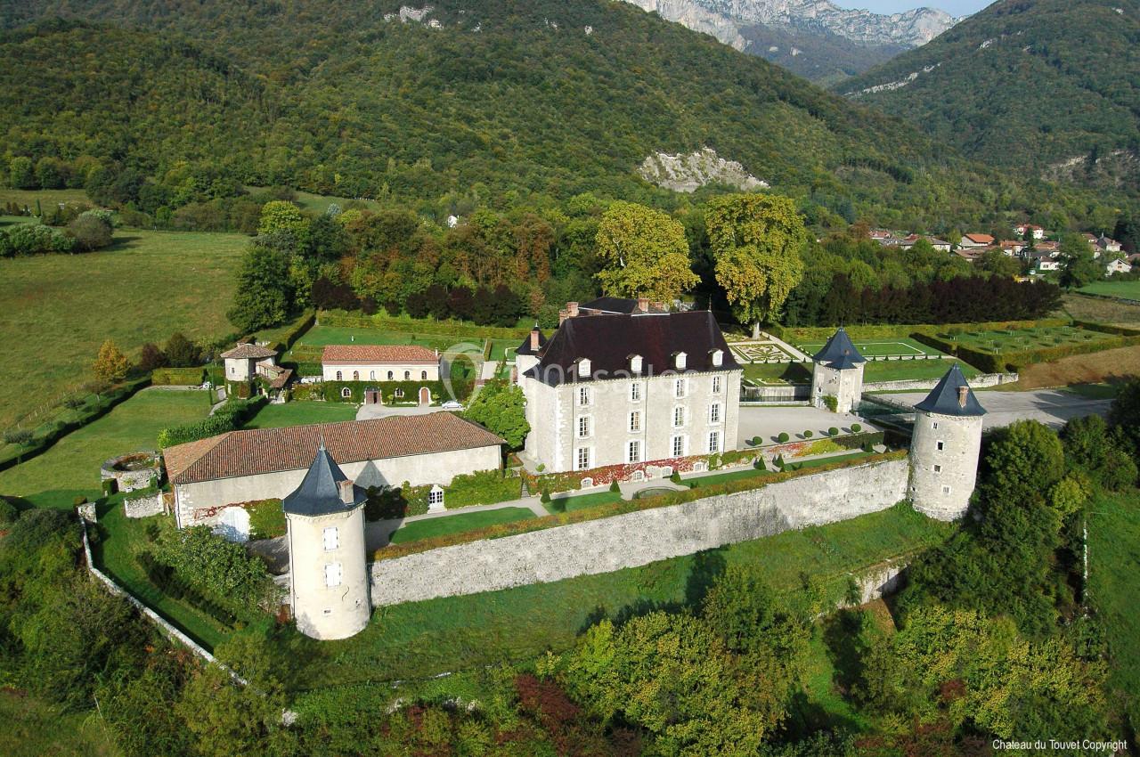 Vue aérienne d'un château entouré de tours, de bâtiments annexes et de verdure, situé dans une vallée montagneuse.