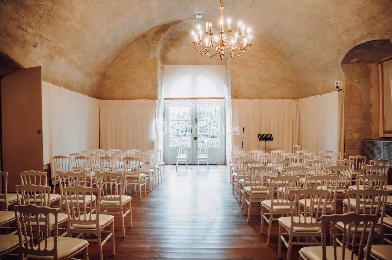 Salle voûtée lumineuse avec des rangées de chaises blanches disposées face à une fenêtre et un lustre central.