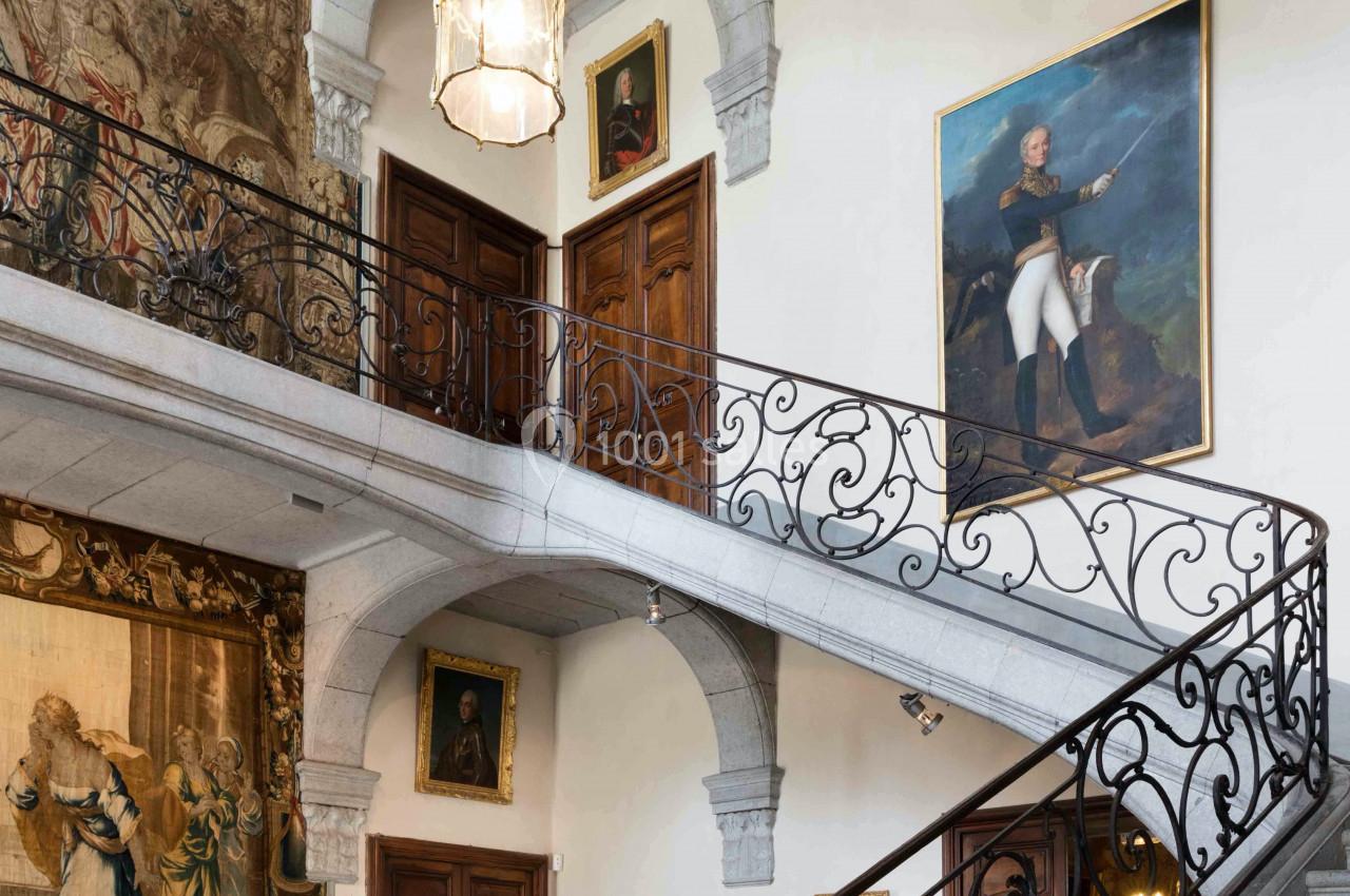 Escalier intérieur en pierre avec balustrades en fer forgé, entouré de tableaux, tapisseries et portes en bois.